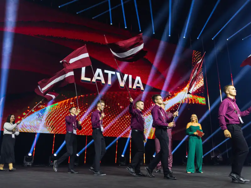 Latvijas komanda ar karogiem iznāk uz skatuves zem uzraksta LATVIA